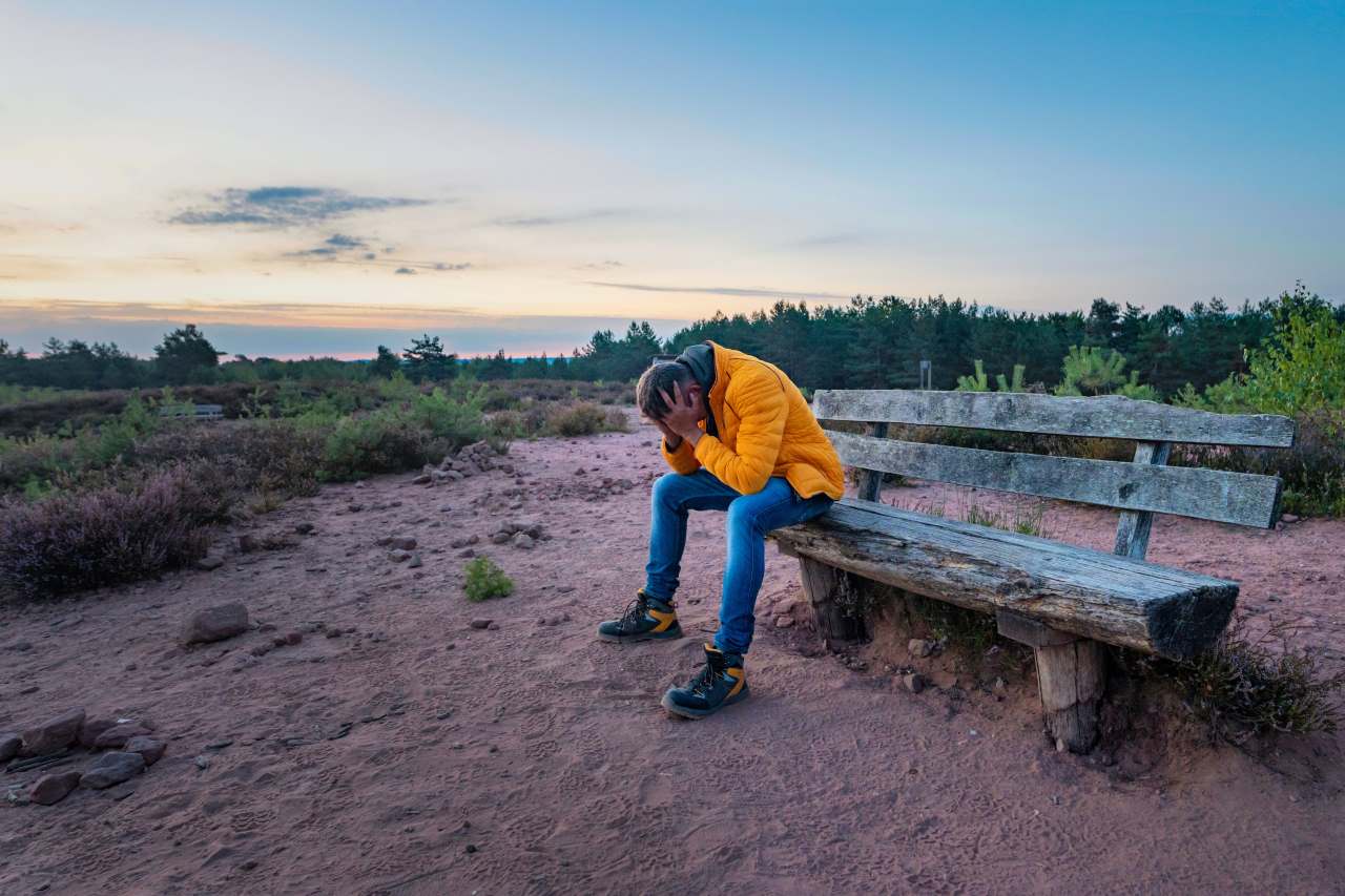 dealing with loneliness, clutching his head in his hands on an old wooden bench on a rural field