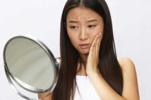 A woman looking in a hand mirror with an expression of concern.